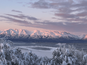 Snow-covered mountains at sunrise, reflecting soft pink hues over a serene river valley below.