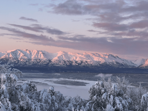 Snow-covered mountains at sunrise, reflecting soft pink hues over a serene river valley below.
