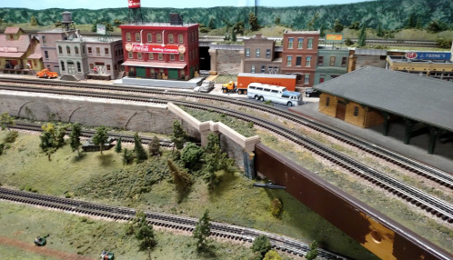 A detailed model train layout featuring buildings, trees, and tracks, with a bus and vehicles on the streets.
