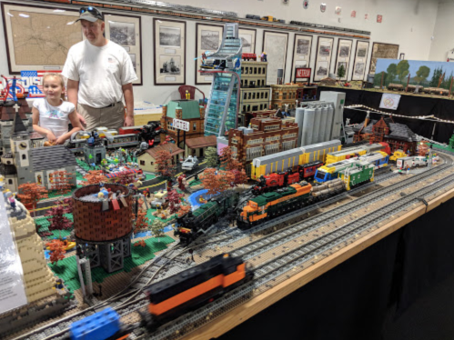A detailed LEGO train display with buildings, trees, and a child and adult observing the scene in the background.