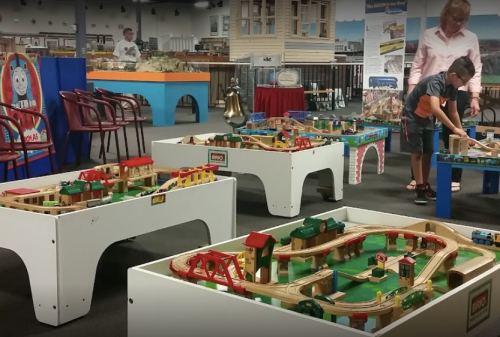 A child plays with wooden train sets on tables in a colorful room filled with model trains and structures.
