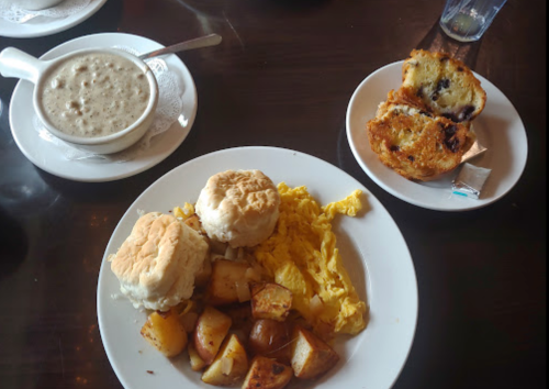 A plate with scrambled eggs, biscuits, roasted potatoes, a bowl of soup, and a muffin on the side.
