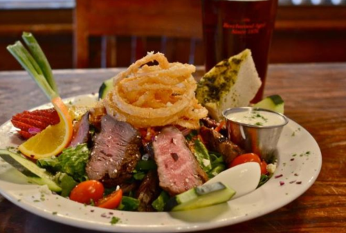 A colorful salad topped with sliced steak, onion rings, and garnished with vegetables, served with a drink in the background.