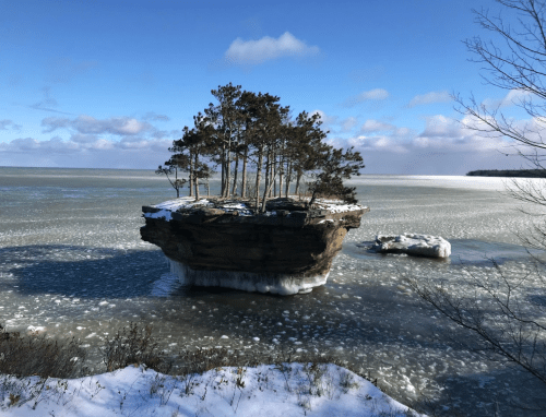 A small island with trees sits atop a frozen lake, surrounded by ice and snow under a clear blue sky.