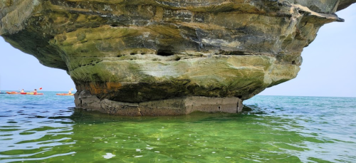 A large rock formation overhanging clear green water, with kayakers visible in the distance.