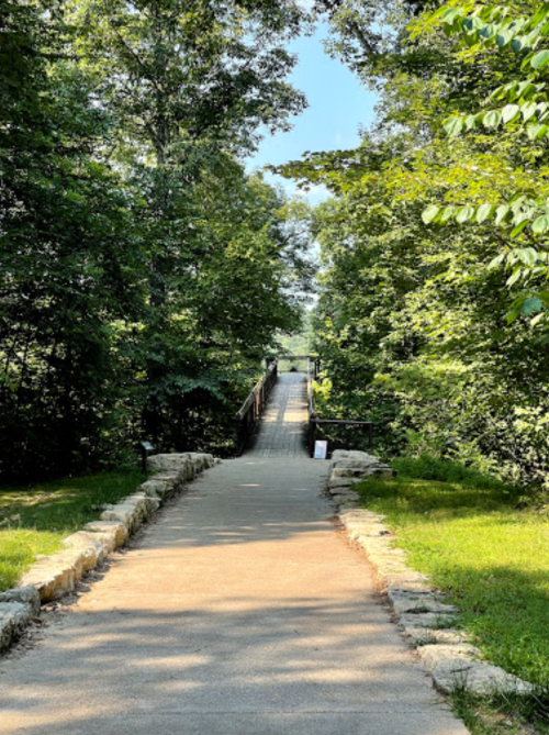 A paved path leads through lush greenery to a wooden bridge in the distance, under a clear blue sky.