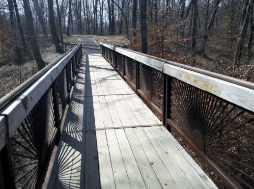 A wooden bridge with decorative railings spans a path through a wooded area, surrounded by bare trees.