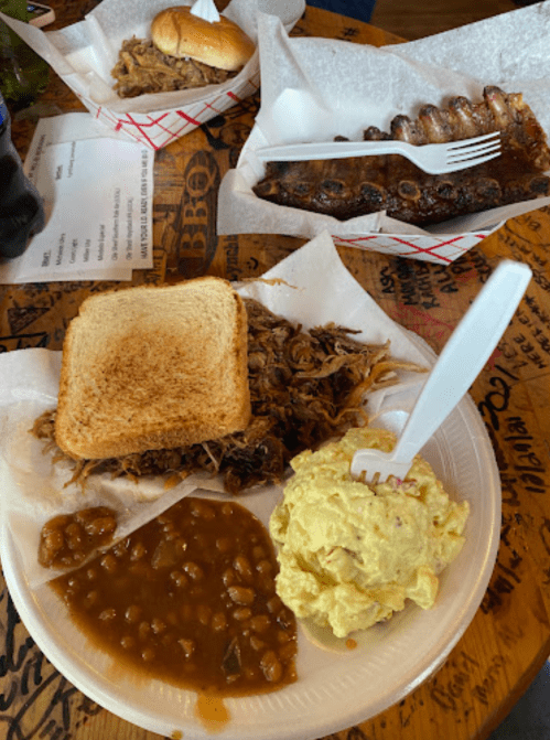 A plate with pulled pork on toast, baked beans, and potato salad, with a side of ribs and a sandwich in the background.