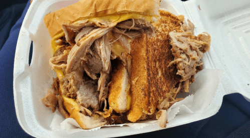 A close-up of a messy sandwich filled with pulled pork and melted cheese, served in a takeout container.