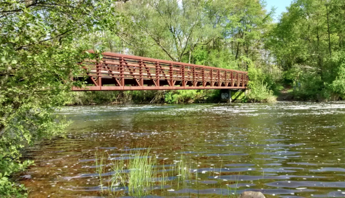A wooden bridge spans a calm river, surrounded by lush greenery and trees under a clear blue sky.