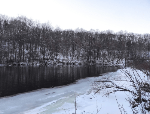 A serene winter landscape featuring a partially frozen river bordered by snow-covered trees.