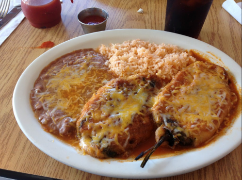 A plate of three cheesy stuffed peppers with rice and refried beans, accompanied by a small cup of sauce.