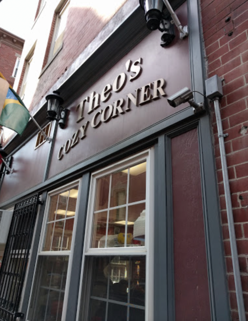 Sign for "Theo's Cozy Corner" on a brick building, featuring large windows and outdoor lighting.