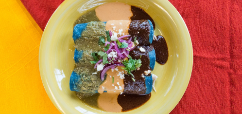 A vibrant plate of blue corn enchiladas topped with green sauce, red sauce, onions, and cilantro on a colorful tablecloth.