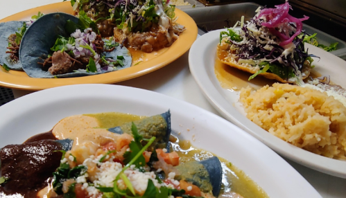 A colorful spread of tacos on blue corn tortillas, topped with fresh greens, alongside rice and a vibrant sauce.