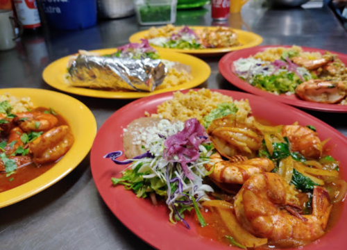 A colorful spread of shrimp dishes on yellow and red plates, featuring rice, salad, and a foil-wrapped burrito.