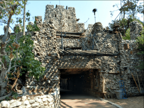 A stone castle-like structure with a wooden bridge and trees surrounding it, set in a sunny outdoor environment.