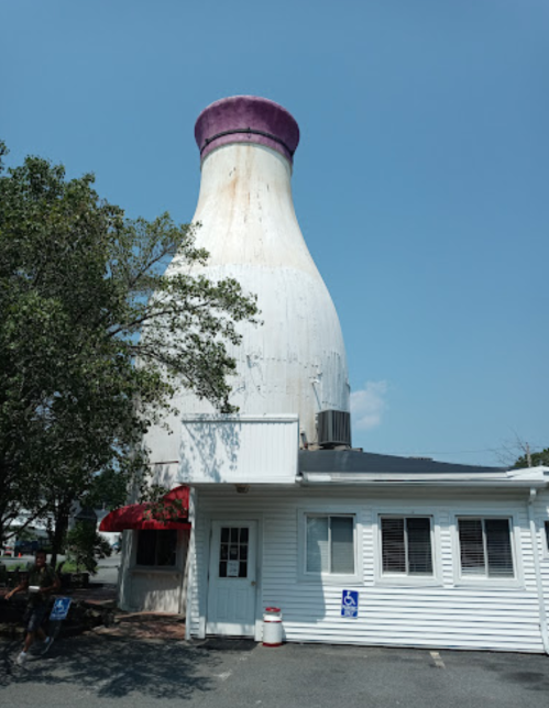 A large, white milk bottle-shaped structure towers over a small building under a clear blue sky.