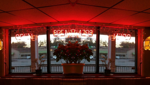A cozy restaurant window with a "BREAKFAST" neon sign, surrounded by festive lights and a potted plant.
