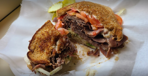 A close-up of a sliced roast beef sandwich with tomatoes, lettuce, and toasted bread, served with pickles.