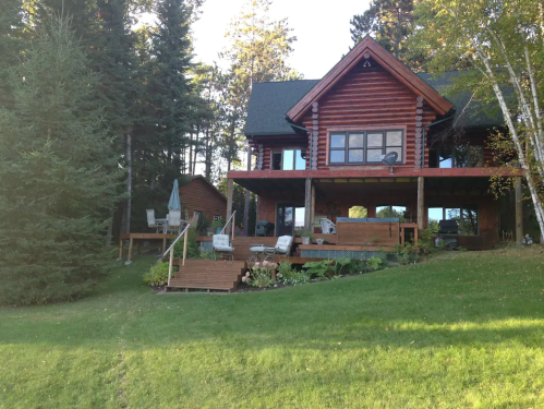 A log cabin home surrounded by trees, featuring a spacious deck and lawn with outdoor seating.