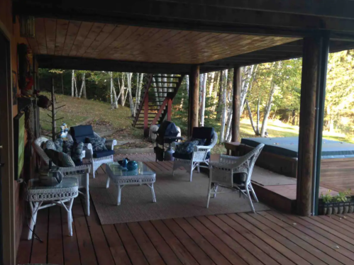Cozy outdoor seating area with wicker chairs, a rug, and a hot tub, surrounded by trees and a wooden deck.