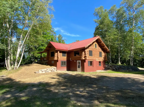 A rustic log cabin with a red roof, surrounded by trees and a grassy area under a clear blue sky.