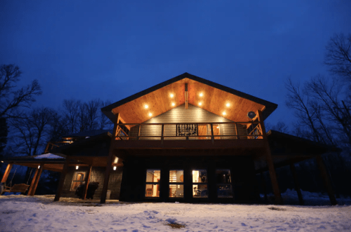 A modern house illuminated at dusk, surrounded by snow and trees, showcasing a warm, inviting atmosphere.