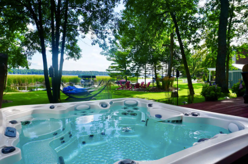 A hot tub on a deck surrounded by trees, with a view of a lake and green lawn furniture in the background.