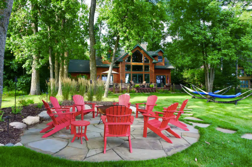 A cozy outdoor seating area with red chairs around a stone patio, surrounded by trees and a house in the background.