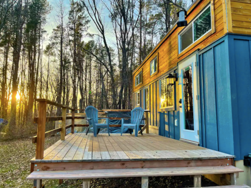 A cozy wooden cabin with a deck, surrounded by trees, illuminated by a warm sunset glow. Two blue chairs are on the deck.