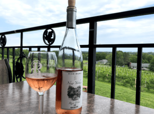 A bottle of rosé wine and a glass sit on a table with a vineyard view in the background under a cloudy sky.