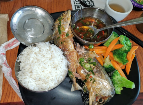 A plate with two grilled fish, rice, steamed broccoli, and carrots, accompanied by a small bowl of dipping sauce.