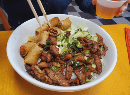 A bowl of stir-fried meat with green onions, cucumber, and spring rolls, served with a drink in the background.