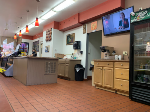 Interior of a casual eatery with a counter, TV on the wall, and a drink fridge, featuring warm lighting and tiled floor.
