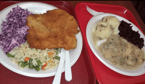 Two plates of food on red trays: one with fried chicken, rice, coleslaw, and mixed vegetables; the other with dumplings and gravy.