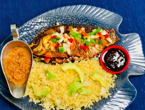 A plate featuring grilled fish topped with vegetables, served with couscous, avocado slices, and two sauces on the side.