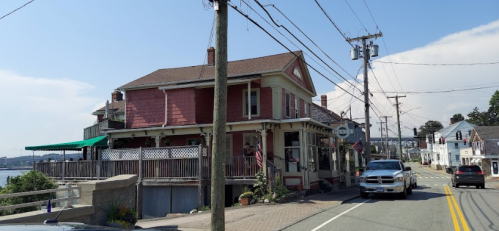 A pink building with a porch sits by the road, with cars passing and power lines overhead, near a waterfront.