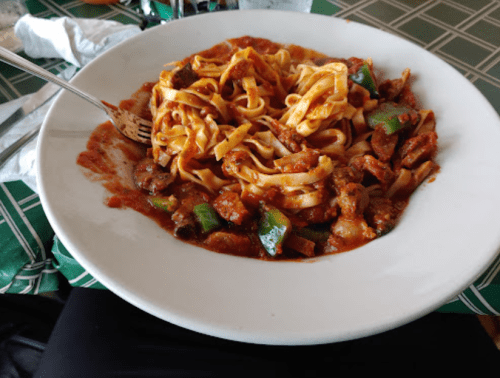 A plate of fettuccine pasta with a rich tomato sauce, green peppers, and pieces of meat, served in a white bowl.