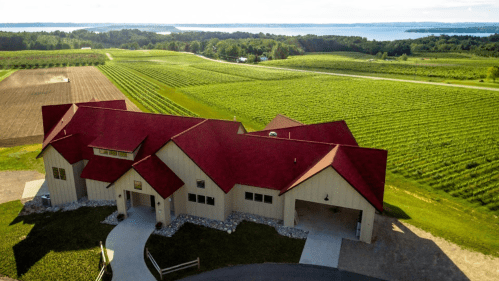 Aerial view of a large house with a red roof, surrounded by lush green fields and a lake in the background.