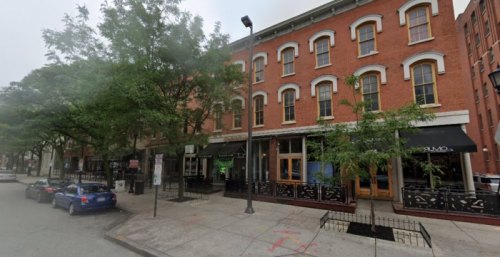 A street view of a brick building with large windows, trees, and parked cars along the sidewalk.