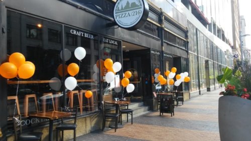 Exterior of a pizza restaurant with orange and white balloons, outdoor seating, and a sign for craft beer.