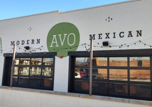 Exterior of AVO Modern Mexican restaurant, featuring a green logo and large windows with string lights.