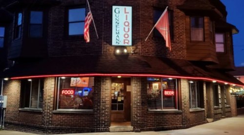 A brick building with a neon sign reading "Gunzelman's Liquor," featuring flags and illuminated windows at dusk.