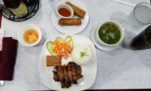A plate of grilled meat with rice, pickled vegetables, spring rolls, and a bowl of soup on a table.