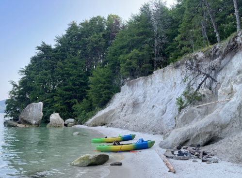 Two colorful kayaks on a sandy beach beside a calm lake, with a rocky shoreline and lush green trees in the background.