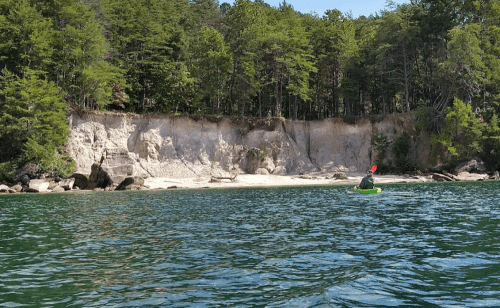 A person kayaking near a sandy beach and steep, tree-lined cliffs on a calm water body.