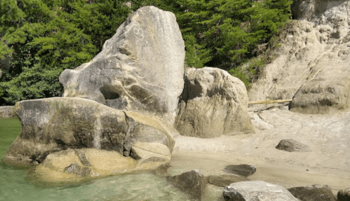 A large rock formation by a sandy beach, surrounded by greenery and calm water under a clear sky.