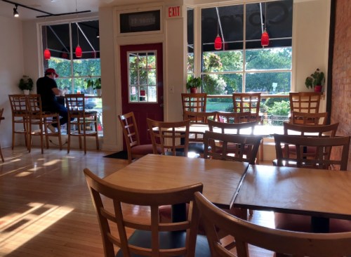 A cozy café interior with wooden tables and chairs, large windows, and a person sitting by the window.