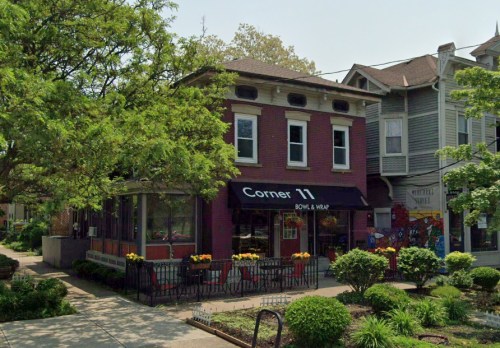 A brick building with a black awning labeled "Corner 11 Bowl & Wrap," surrounded by greenery and colorful flowers.